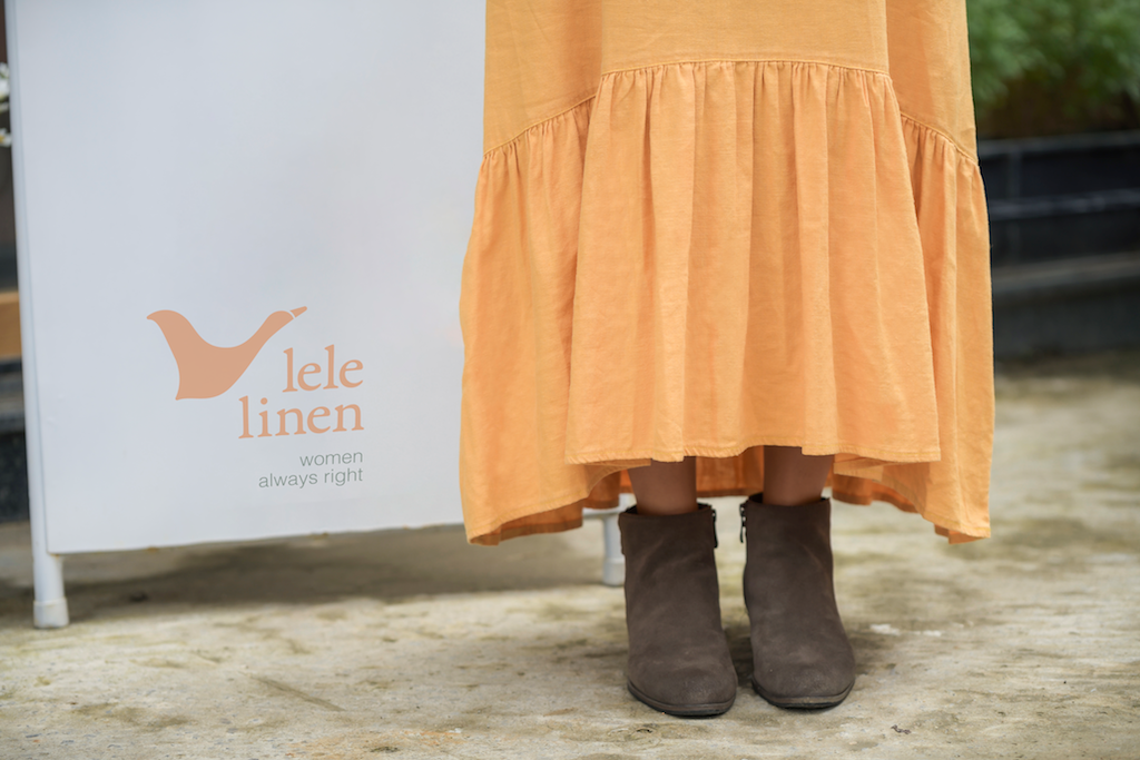 Lele Linen studio sign beside a customer wearing an apricot linen dress with ankle boots.